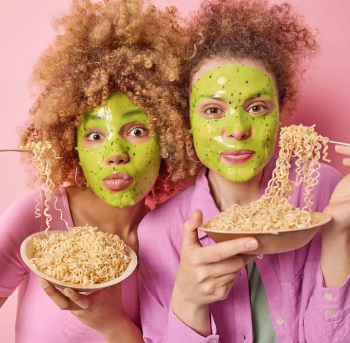 Deux femmes avec un masque de soin vert sur le visage mangeant des nouilles, illustrant la double expertise en analyse sensorielle agroalimentaire et cosmétique de Techni'Sens.
